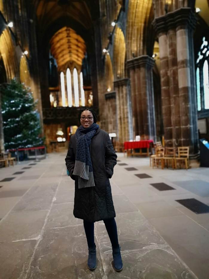 A beautiful, tall lady wearing a black winter jacket with a navy-blue muffler wrapped around her neck, standing inside an intricately designed cathedral.